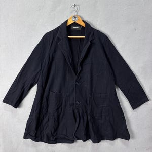 May include: A black blazer with a two-button fastening and two front pockets. The blazer features long sleeves and a distinctive flared hem. The garment is suspended on a wooden hanger against a grey backdrop.