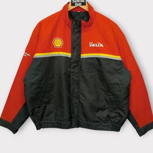 May include: Red and black Shell Helix jacket with a stand-up collar. The jacket features the Shell logo and Helix branding on the chest. A yellow and gray stripe runs across the chest. The jacket is hanging on a hanger.