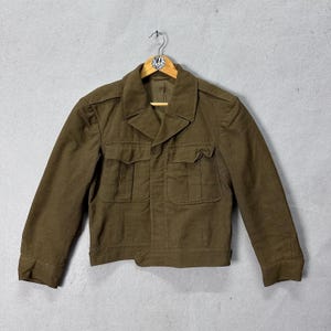 May include: Olive green jacket with two chest pockets and two lower pockets. The jacket has a collared neckline and button closure. It is hanging on a wooden hanger against a gray background. The jacket appears to be made of a durable fabric.
