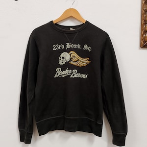 Può includere: Felpa girocollo nera con la scritta "23rd Bomb. Sq." sopra un teschio con le ali e le parole "Bomber Barons" sotto. La grafica è nei colori crema, oro e arancione. La felpa è appesa a una gruccia di legno.