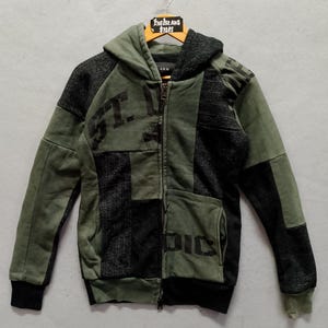 May include: Green and black patchwork zip-up hoodie with the text "ST. LUC" and "PIC" printed on the front. The hoodie has a hood, front pockets, and black ribbed cuffs and hem. The Island Store is printed on the hanger.