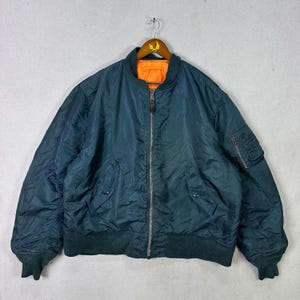 Puede incluir: Una chaqueta bomber verde azulado con cuello y puños verde oscuro e interior naranja. La chaqueta tiene cierre de cremallera, dos bolsillos delanteros y un pequeño bolsillo en la manga izquierda. Cuelga de una percha de madera.