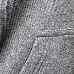 May include: Close-up of a gray hoodie pocket. The pocket is rectangular and stitched to the main fabric. The fabric is a medium gray color and has a textured appearance. There is a small tear in the fabric near the pocket.