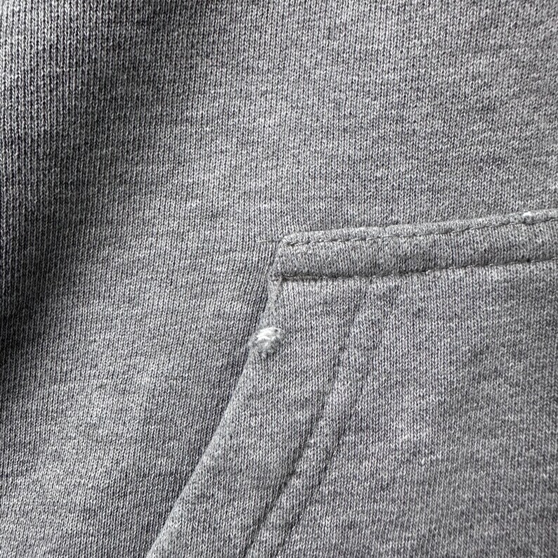 May include: Close-up of a gray hoodie pocket. The pocket is rectangular and stitched to the main fabric. The fabric is a medium gray color and has a textured appearance. There is a small tear in the fabric near the pocket.
