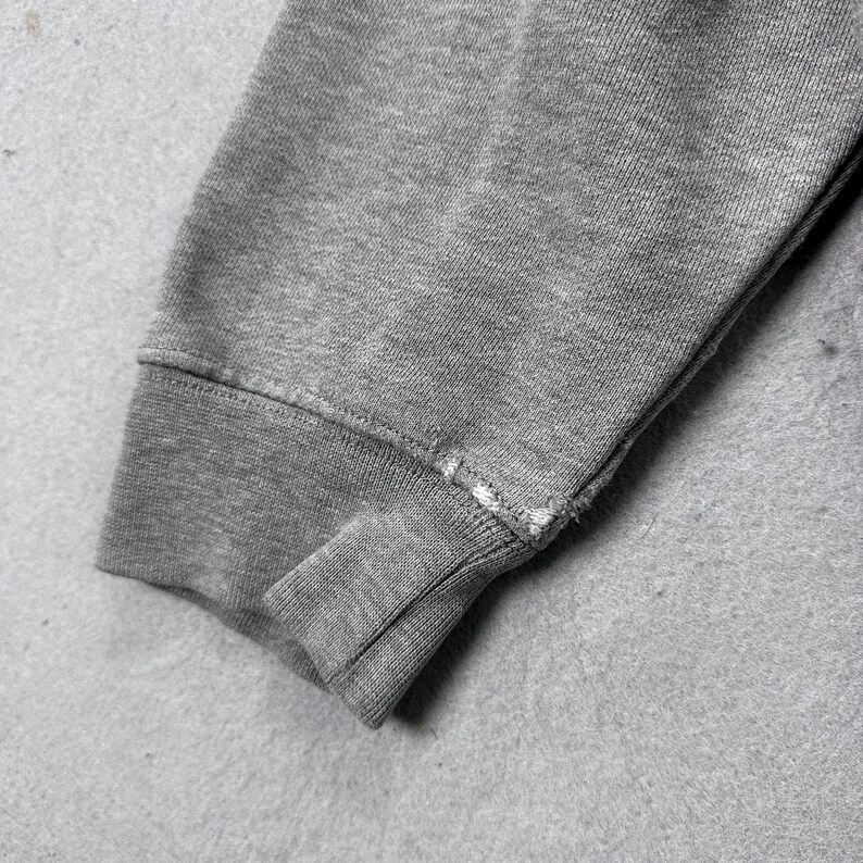 May include: Close-up of a gray sweatshirt sleeve, showing the ribbed cuff and a portion of the arm. The fabric appears to be a soft, knit material. The cuff has a seam with visible stitching.