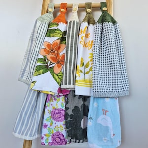 May include: A wooden ladder displays a collection of hanging kitchen towels. The towels feature various patterns, including floral, striped, and gingham designs. Each towel has a crocheted top with a button closure. The colour palette includes green, orange, white, and blue.