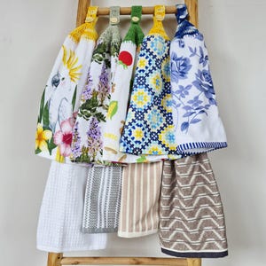 May include: A collection of hanging kitchen towels featuring various patterns including a white cockatoo, floral prints, geometric designs, and stripes. Each towel has a crocheted top with a button closure, designed to hang from a wooden rack.