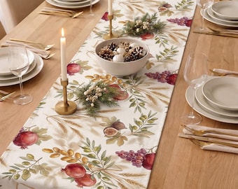 May include: A cream-colored table runner featuring a botanical print of pomegranates, grapes, figs, wheat stalks, and olive branches. The runner is decorated with two gold-toned candle holders with white taper candles and a central bowl of pinecones.