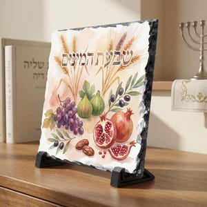May include: A decorative slate plaque featuring a watercolor illustration of wheat, grapes, figs, olives, pomegranates, and dates. The Hebrew text "Sheva HaMinim" is displayed at the top. The plaque is supported by a black stand and placed on a wooden surface.