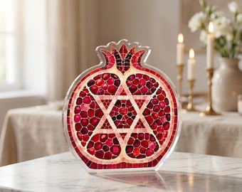 May include: A pomegranate-shaped decorative object made of clear acrylic with a mosaic-style interior. The design features deep red and pink segments resembling seeds, with a prominent white Star of David centered within the fruit's silhouette.