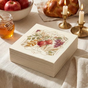 May include: A stack of square paper napkins featuring a decorative border with a watercolor illustration of pomegranates, grapes, and wheat. The text "CHAG SAMEACH" and "Cohen Family" is printed in the center. The napkins are off-white.