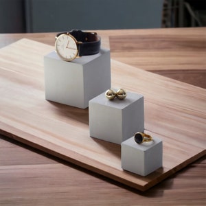 Puede incluir: Tres expositores cuadrados grises de diferentes tamaños sobre una superficie de madera. Un reloj de pulsera se muestra en el expositor más grande, dos esferas doradas están en el expositor del medio y un anillo con una piedra negra está en el expositor más pequeño.