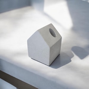 Peut inclure: Un petit porte-crayons en béton blanc en forme de maison avec un trou rond sur le dessus.