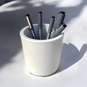 Op de afbeelding: Een witte betonnen potloodhouder met verschillende zwarte pennen erin. De houder is cilindrisch en heeft een glad, getextureerd oppervlak.