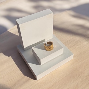 Peut inclure: Une bague en or avec une surface texturée est posée sur un présentoir blanc. Le présentoir est composé de deux carrés empilés.