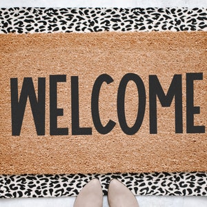 May include: A brown coir doormat with a black leopard print border. The doormat has the word "WELCOME" printed in black.