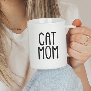 K&ouml;nnte beinhalten: Eine wei&szlig;e Keramiktasse mit schwarzem Text, der "CAT MOM" lautet.
