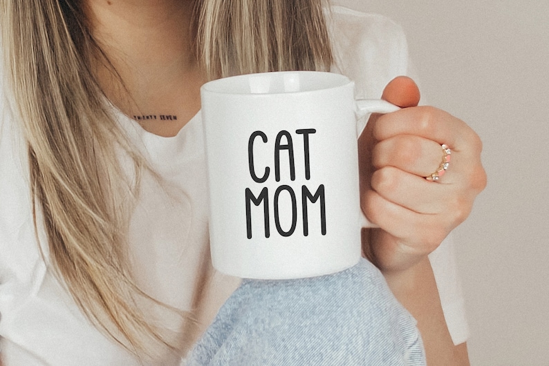 K&ouml;nnte beinhalten: Eine wei&szlig;e Keramiktasse mit schwarzem Text, der "CAT MOM" lautet.