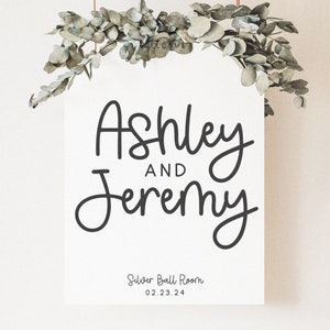 Peut inclure: Une pancarte blanche avec une calligraphie noire qui dit "Ashley et Jeremy" avec le texte "Silver Ball Room 02.23.24" en dessous. La pancarte est suspendue &agrave; une guirlande de feuilles d'eucalyptus.