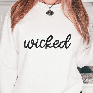 Puede incluir: Sudadera blanca con la palabra "wicked" en escritura negra. La imagen tiene un tema de Halloween, con decoraciones de murci&eacute;lagos y una taza con forma de fantasma.