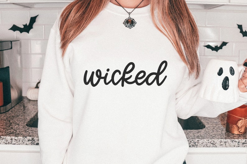 Puede incluir: Sudadera blanca con la palabra "wicked" en escritura negra. La imagen tiene un tema de Halloween, con decoraciones de murci&eacute;lagos y una taza con forma de fantasma.