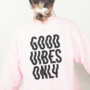 Op de afbeelding: Roze sweatshirt met de tekst "GOOD VIBES ONLY" in het zwart gedrukt op de achterkant.