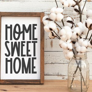 May include: A framed sign with the text "Home Sweet Home" in black lettering on a white background. The sign is sitting on a wooden surface with a vase of cotton flowers in the background.