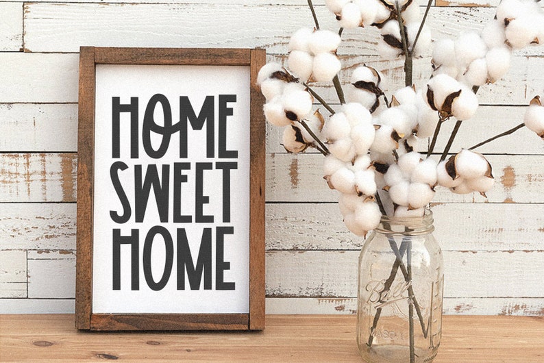 May include: A framed sign with the text "Home Sweet Home" in black lettering on a white background. The sign is sitting on a wooden surface with a vase of cotton flowers in the background.