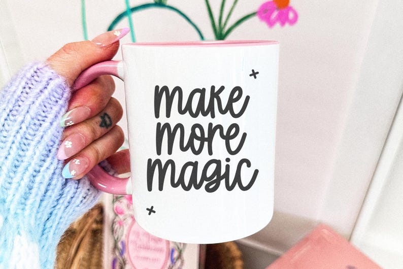 Op de afbeelding: Witte keramische mok met een roze rand en handvat. De mok heeft zwarte tekst met de tekst "make more magic".