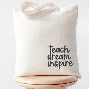 May include: A white canvas tote bag with black text that reads "teach dream inspire".