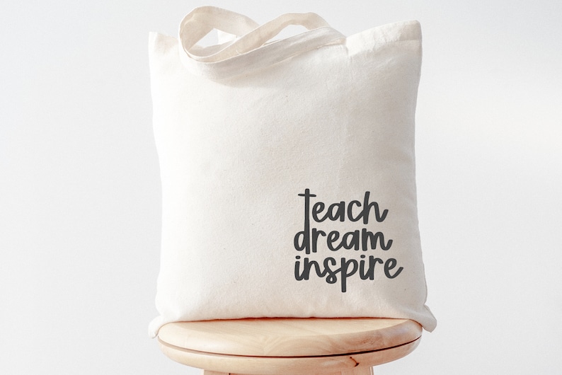 May include: A white canvas tote bag with black text that reads "teach dream inspire".
