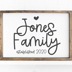 Peut inclure: Une pancarte en bois blanc avec un cadre brun, sur laquelle est inscrit le texte "Jones Family established 2020" en &eacute;criture cursive noire avec un petit c&oelig;ur au-dessus du nom.