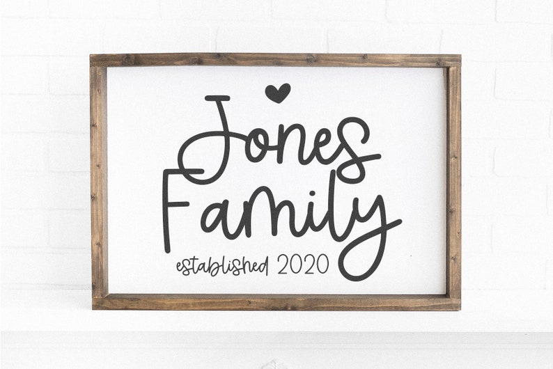 Peut inclure: Une pancarte en bois blanc avec un cadre brun, sur laquelle est inscrit le texte "Jones Family established 2020" en &eacute;criture cursive noire avec un petit c&oelig;ur au-dessus du nom.