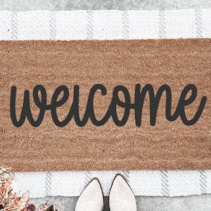 Op de afbeelding: Een bruine kokosmat met het woord "welcome" in zwarte cursieve letters.