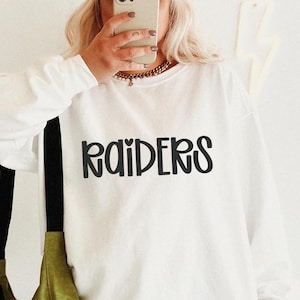 May include: White long-sleeve sweatshirt with the word "RAIDERS" in black, stylized lettering. The sweatshirt is worn by a person holding a phone. A black strap and olive green bag are visible.