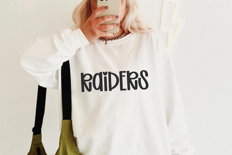 May include: White long-sleeve sweatshirt with the word "RAIDERS" in black, stylized lettering. The sweatshirt is worn by a person holding a phone. A black strap and olive green bag are visible.