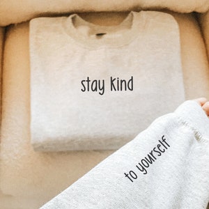 K&ouml;nnte beinhalten: Ein grauer Pullover mit dem Aufdruck "stay kind to yourself". Der Pullover ist gefaltet und liegt auf einer wei&szlig;en, flauschigen Oberfl&auml;che.