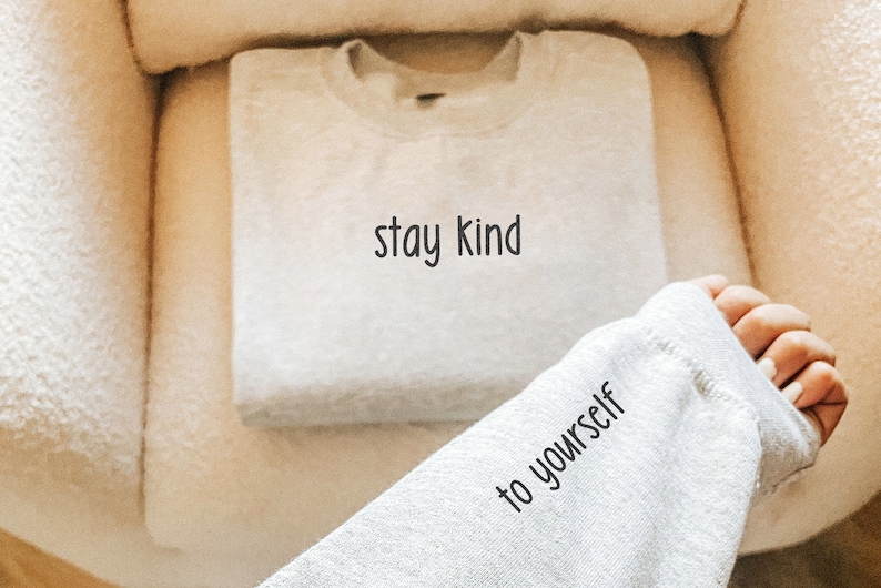 K&ouml;nnte beinhalten: Ein grauer Pullover mit dem Aufdruck "stay kind to yourself". Der Pullover ist gefaltet und liegt auf einer wei&szlig;en, flauschigen Oberfl&auml;che.