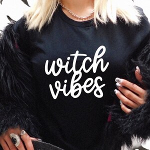 Puede incluir: Camiseta negra con las palabras "witch vibes" en escritura blanca. La persona lleva una chaqueta de piel sint&eacute;tica negra. El fondo tiene un patr&oacute;n de formas de hojas negras.