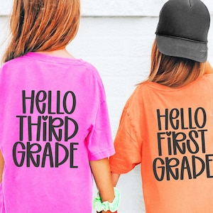 May include: Two children wearing pink and orange t-shirts with the text "HELLO THIRD GRADE" and "HELLO FIRST GRADE" in black. One child wears a black cap. The children are holding hands.