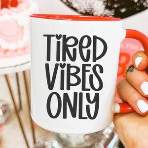 May include: White ceramic mug with a red interior and handle, featuring the text "TIRED VIBES ONLY" in black. The mug is held by a hand with white nail polish and gold rings. A cake and disco balls are in the background.