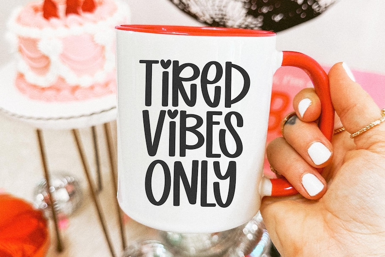 May include: White ceramic mug with a red interior and handle, featuring the text "TIRED VIBES ONLY" in black. The mug is held by a hand with white nail polish and gold rings. A cake and disco balls are in the background.