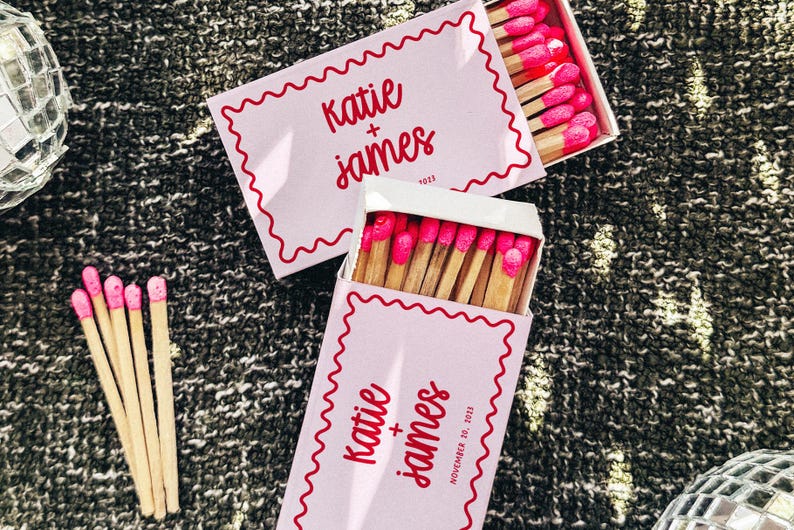 Op de afbeelding: Twee roze luciferdoosjes met een witte rand en rode golvende lijnen. Het bovenste doosje heeft de tekst "Katie + James" in rood. Het onderste doosje heeft de tekst "Katie + James" in rood en de datum "November 12, 2021".