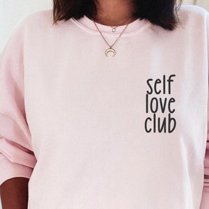 K&ouml;nnte beinhalten: Ein rosa Sweatshirt mit dem Aufdruck "self love club" auf der Vorderseite.