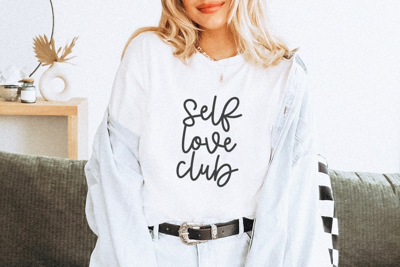 Peut inclure: Un t-shirt blanc avec le texte "Self love club" &eacute;crit en cursive noire.