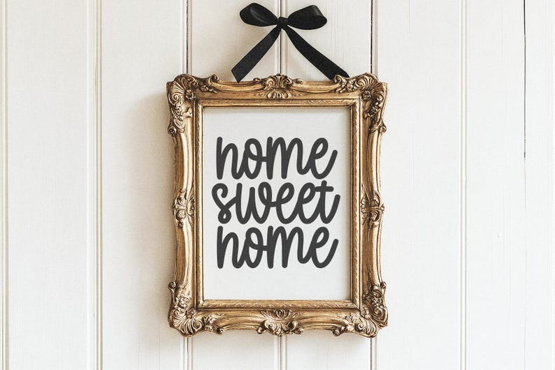 Op de afbeelding: Een gouden lijst met een witte achtergrond en zwarte tekst die "home sweet home" zegt, hangt aan een witte muur.
