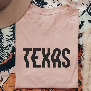 Op de afbeelding: Een roze T-shirt met het woord "TEXAS" in zwarte blokletters.