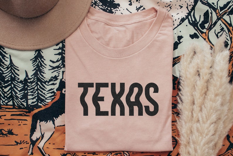 Op de afbeelding: Een roze T-shirt met het woord "TEXAS" in zwarte blokletters.