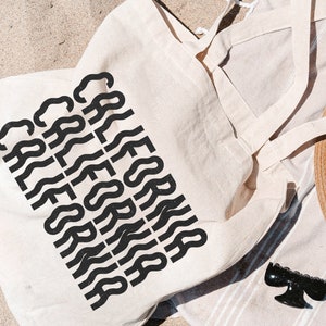 Op de afbeelding: Een witte canvas draagtas met zwarte tekst die meerdere keren "CALIFORNIA" herhaalt in een golvend lettertype. De tas ligt op een gestreepte handdoek met een strohoed en een zwarte haarspeld.