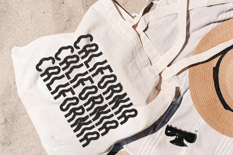 Op de afbeelding: Een witte canvas draagtas met zwarte tekst die meerdere keren "CALIFORNIA" herhaalt in een golvend lettertype. De tas ligt op een gestreepte handdoek met een strohoed en een zwarte haarspeld.